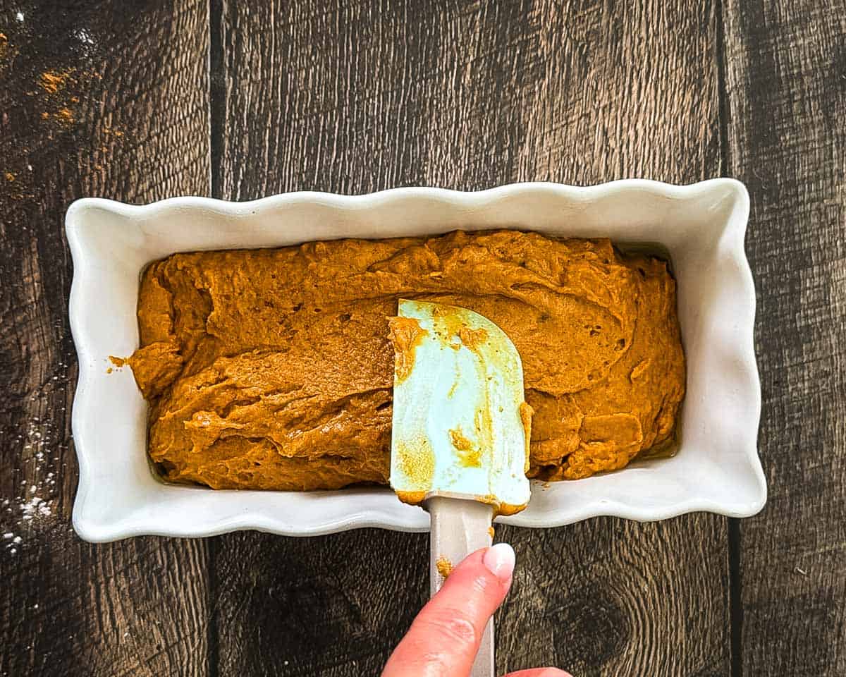 Sourdough discard pumpkin bread batter being spread evenly in a white loaf pan with a spatula.