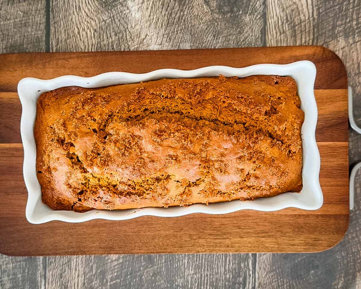 Freshly baked sourdough discard pumpkin bread with a golden brown, cracked crust and demerara sugar topping in a white loaf pan.