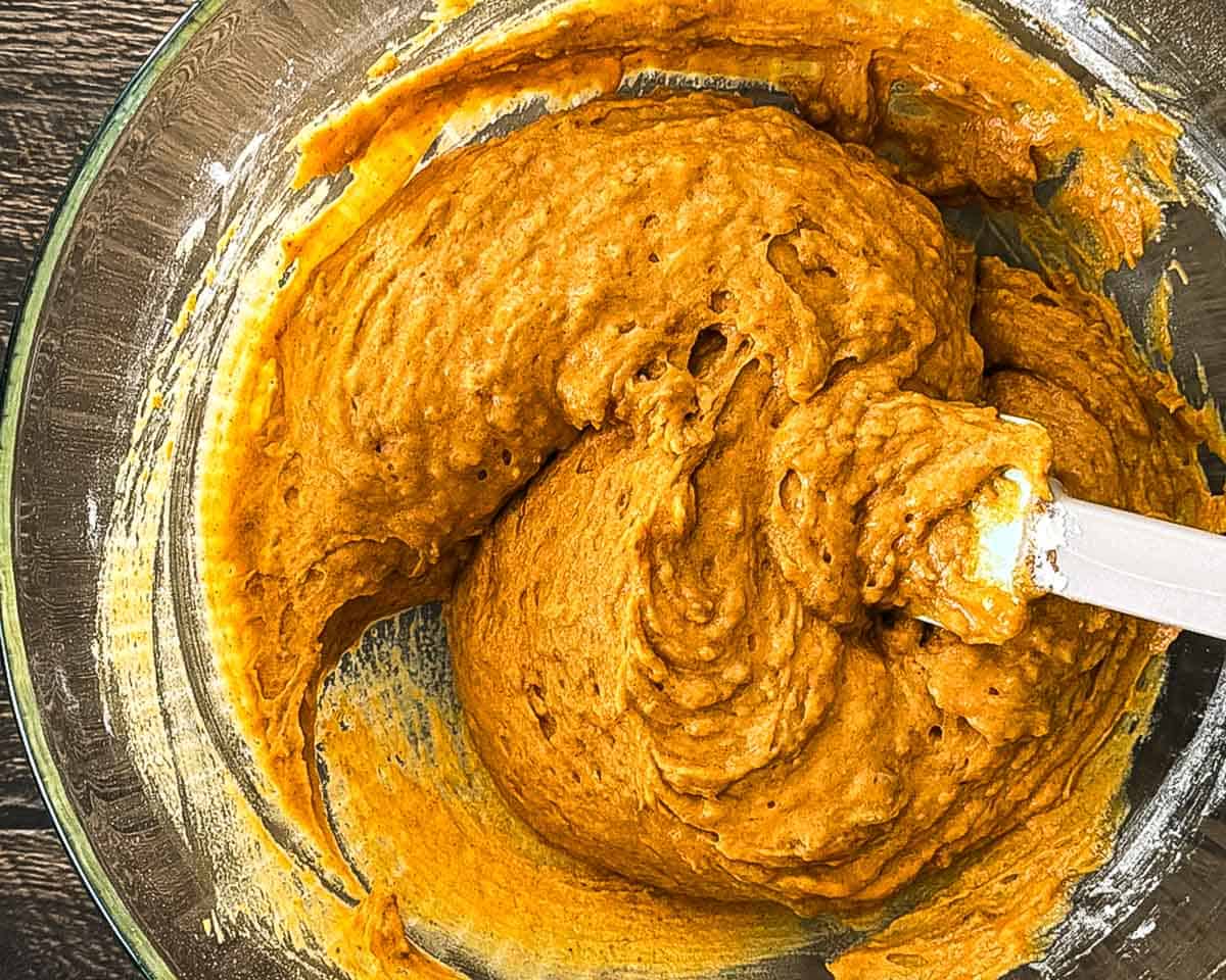 Thick orange sourdough discard pumpkin bread batter being folded together with a spatula in a glass mixing bowl.